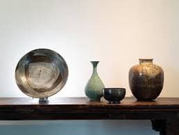 A photograph of four ceramic vessels arranged on a wooden table against a plain white wall. From left to right, there is a large, round, metallic-looking bowl on a stand, a tall green vase, a small dark bowl, and a large brown jar.