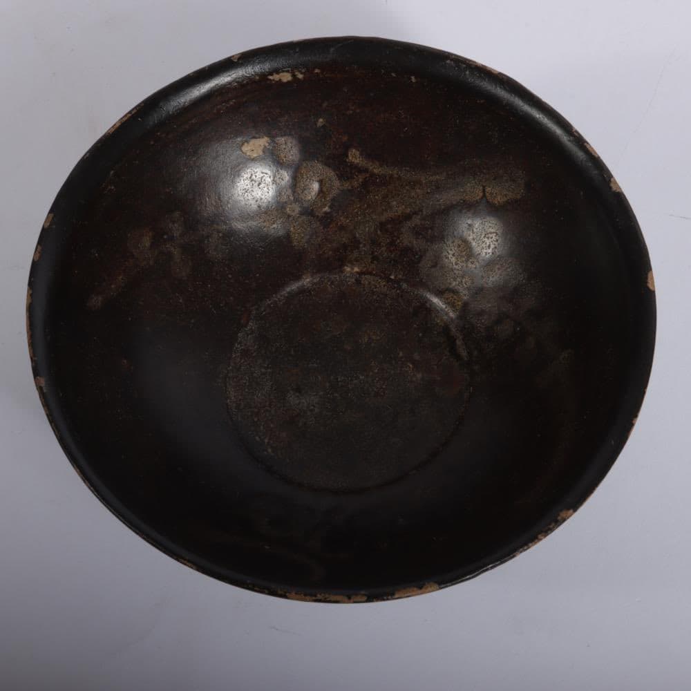 A top-down view of a dark, round bowl with a glossy black glaze and visible wear or residue on its interior surface. The bowl is set against a plain, light-colored background.