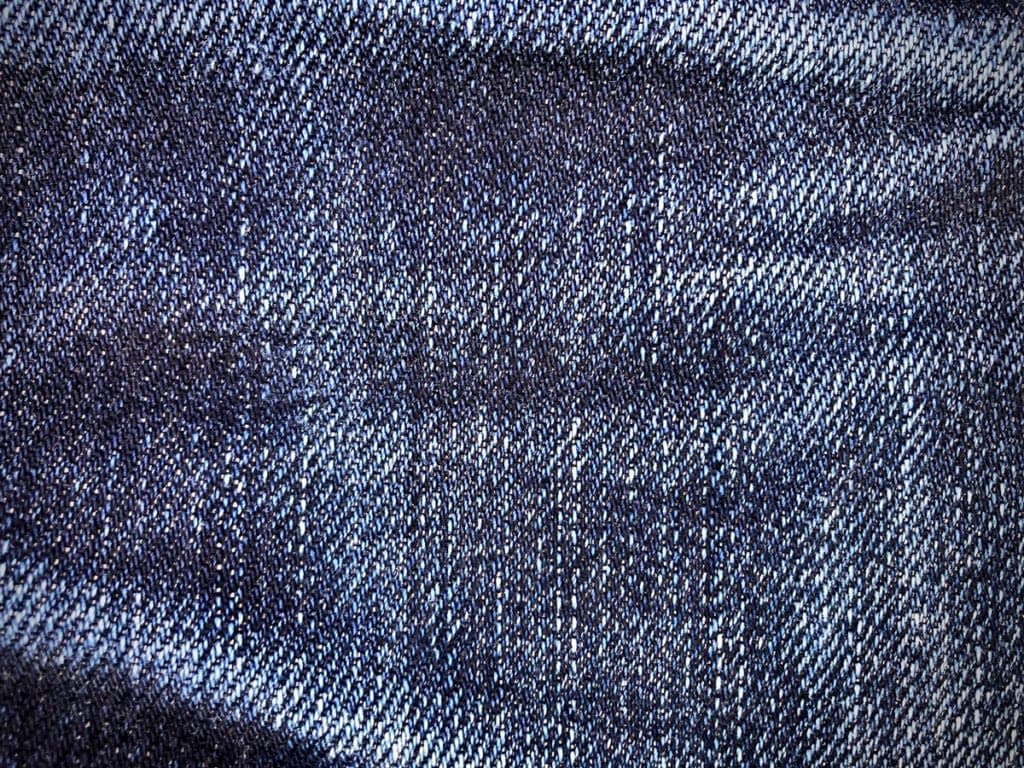 A close-up photograph of a blue denim fabric, showing its textured weave and varying shades of blue. The image focuses on the material's surface, highlighting the diagonal twill pattern characteristic of denim.