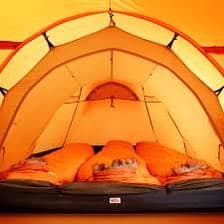 A view from inside a tent looking out through the front opening, showing the interior space with sleeping bags and a sleeping pad. The tent is illuminated with a warm, orange glow, likely from an internal light source or the setting sun.
