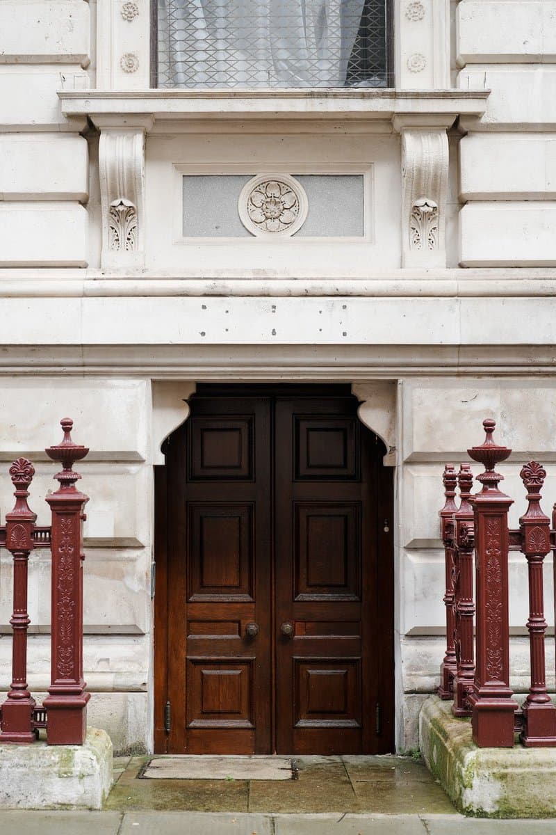 A photograph of an ornate, dark wooden double door set into a white stone building facade, flanked by decorative red-painted iron railings. Above the door is a carved stone panel with a circular floral motif and a window with a leaded glass pattern.
