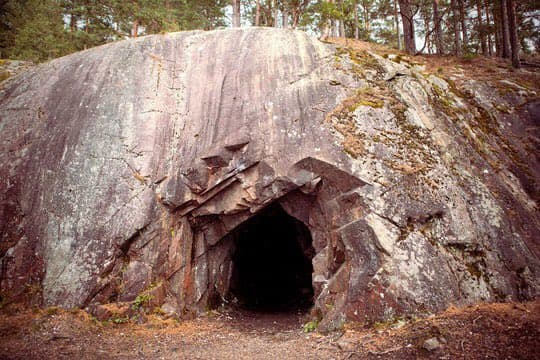 A large, weathered rock formation in a forest setting features a dark, cave-like opening at its base, surrounded by trees and forest floor.