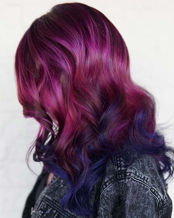 A person shown from the back and side, displaying long, wavy hair dyed in a vibrant gradient from purple and magenta at the top to deep blue at the ends. The person is wearing a dark, textured jacket.