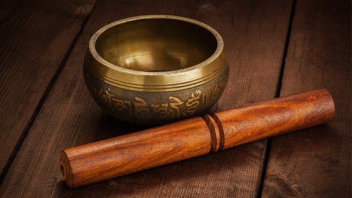 A brass Tibetan singing bowl with engraved script sits on a dark wooden surface next to a wooden mallet.