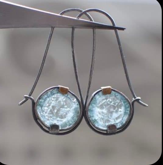 A pair of handmade earrings featuring round, light-blue gemstones set in silver wire, held up by a pair of pliers against a blurred background.
