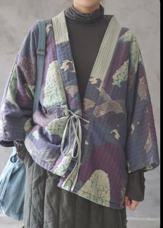 A person models a loose-fitting, patterned kimono-style jacket in shades of purple and blue over a dark turtleneck and trousers, carrying a denim shoulder bag.
