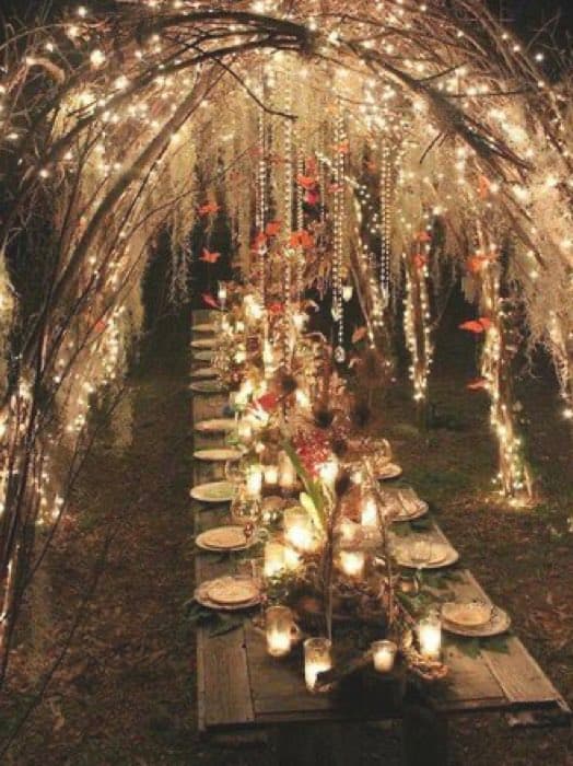 A long outdoor dining table is set with plates, glasses, and numerous lit candles under a rustic archway covered in string lights and hanging ornaments. The scene is illuminated by the warm glow of the lights and candles, creating a romantic and festive atmosphere in a wooded area at night.