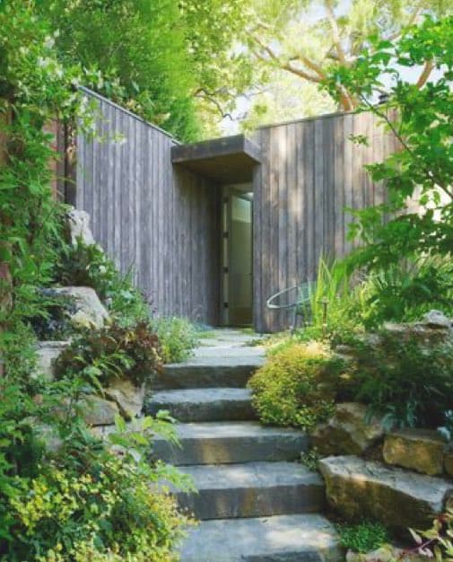 A stone pathway leads up to a modern wooden structure surrounded by lush greenery and foliage. The scene features a minimalist architectural design with vertical wood siding and an open doorway, framed by trees and plants.