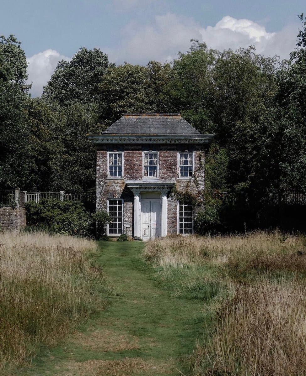 A small, two-story brick cottage with a white door and window frames stands in a field of tall, dry grass, with a mowed path leading to its entrance and a dense line of trees in the background.