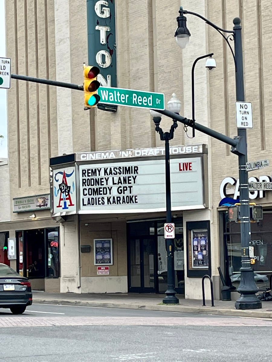 A street-level photograph of the Cinema on Draft House theater, featuring a marquee that lists "COMEDY GPT" among other acts. The scene includes a traffic light, a street sign for Walter Reed Dr, and a vertical sign for the Gorton's restaurant.