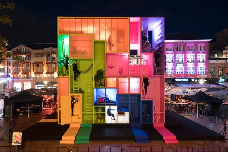 A nighttime photograph of a large, colorful, multi-story art installation resembling a house with transparent rooms and ladders, illuminated with vibrant neon lights in a public square. People are visible inside and climbing the structure, while surrounding buildings and tents are lit in the background.