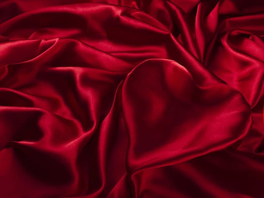 A close-up photograph of a rich, deep red satin fabric with soft, flowing folds and a glossy sheen.