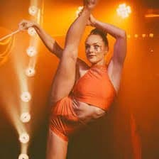 A woman in an orange two-piece outfit performs a high leg split on a stage with warm orange lighting and a string of bright bulbs in the background. She is holding a thin white ribbon or cord in her raised hand.
