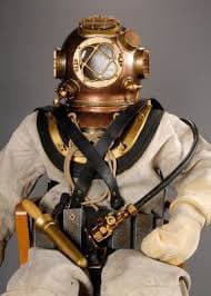 A person is dressed in a vintage deep-sea diving suit, featuring a large brass helmet with a circular glass faceplate and a white canvas suit. The diver is equipped with a harness and a brass air hose connected to a belt-mounted apparatus.