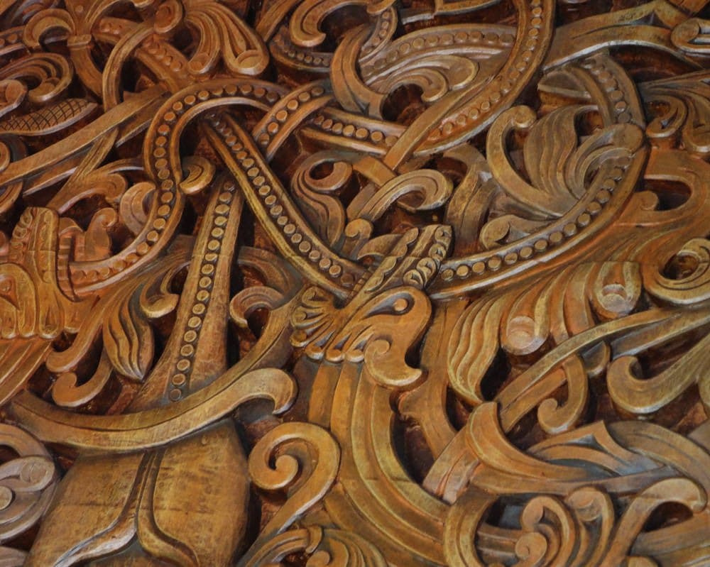A close-up photograph of intricate, swirling wooden carvings in warm brown and gold tones, featuring detailed scrollwork and beaded patterns. The image showcases the texture and depth of the hand-carved woodwork.