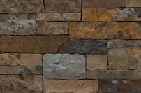 A close-up view of a textured stone wall made of rectangular blocks in various earth tones, including brown, tan, gray, and rust. The stones are arranged in a stacked pattern with visible mortar lines. The image has a natural, rustic appearance.