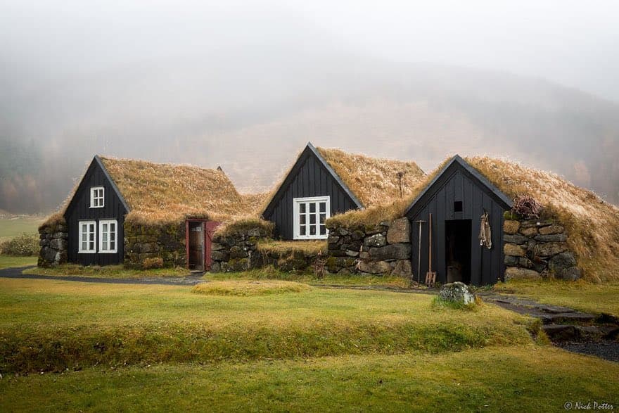 A landscape photograph showing three traditional black wooden buildings with grass-covered roofs and stone foundations, set in a misty, green field.