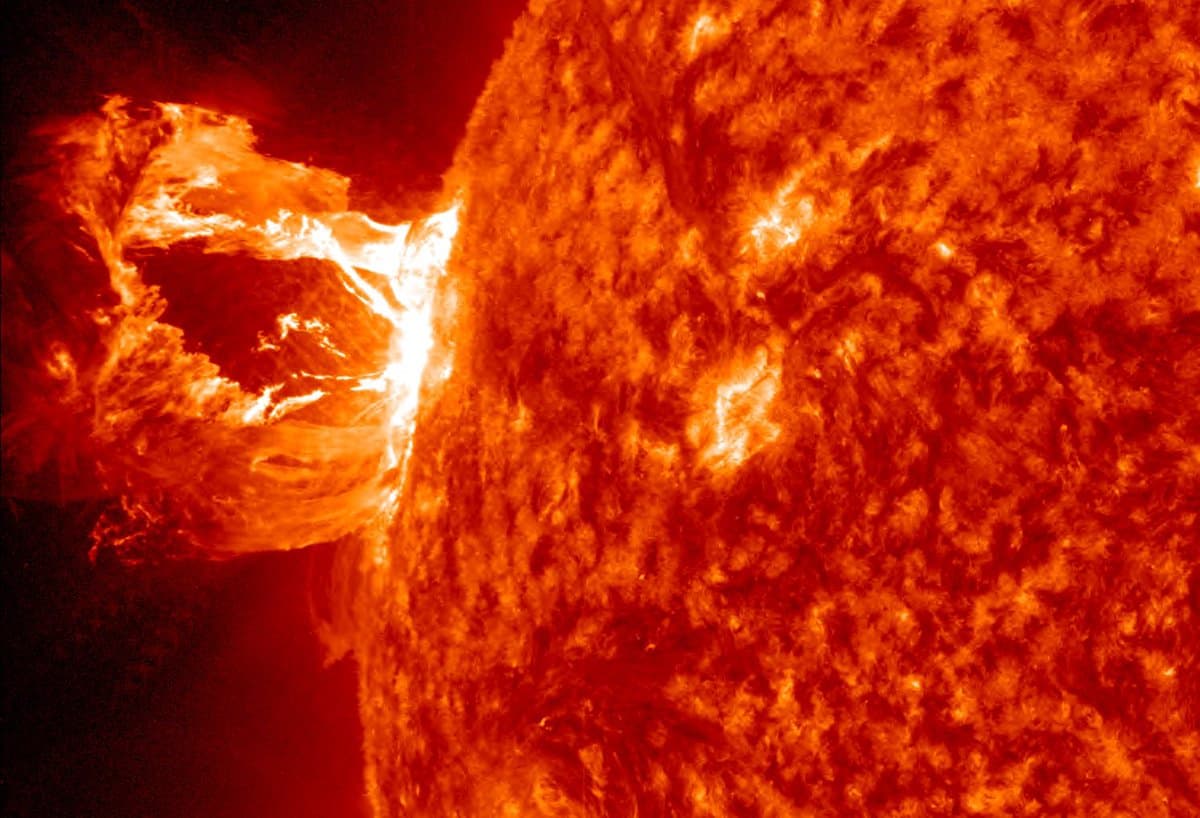 A close-up, high-contrast image of the Sun's surface in fiery orange and red tones, showing a massive solar prominence erupting from the left edge of the star. The image captures the turbulent, textured surface and the bright, looping plasma of the eruption against the dark background of space.