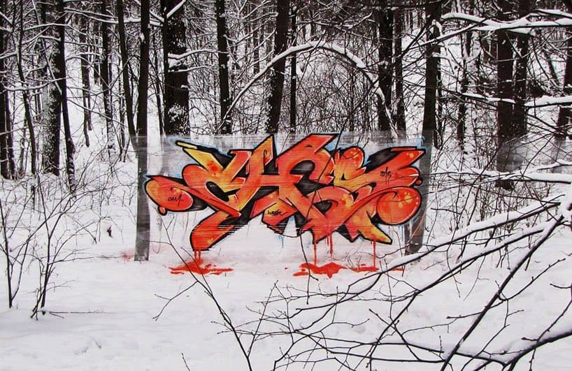 A photograph of a snow-covered forest with a large, colorful graffiti tag painted on a wooden structure in the center. The tag features stylized orange and yellow lettering with black outlines and drips of paint on the snow below.