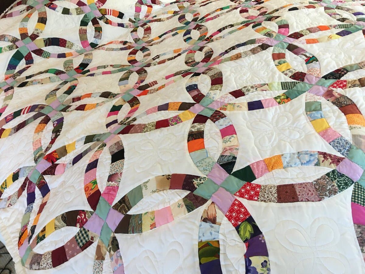 A close-up photograph of a colorful patchwork quilt featuring a repeating pattern of curved, multicolored fabric strips on a white background. The quilt displays intricate stitching and various floral and geometric fabric prints.