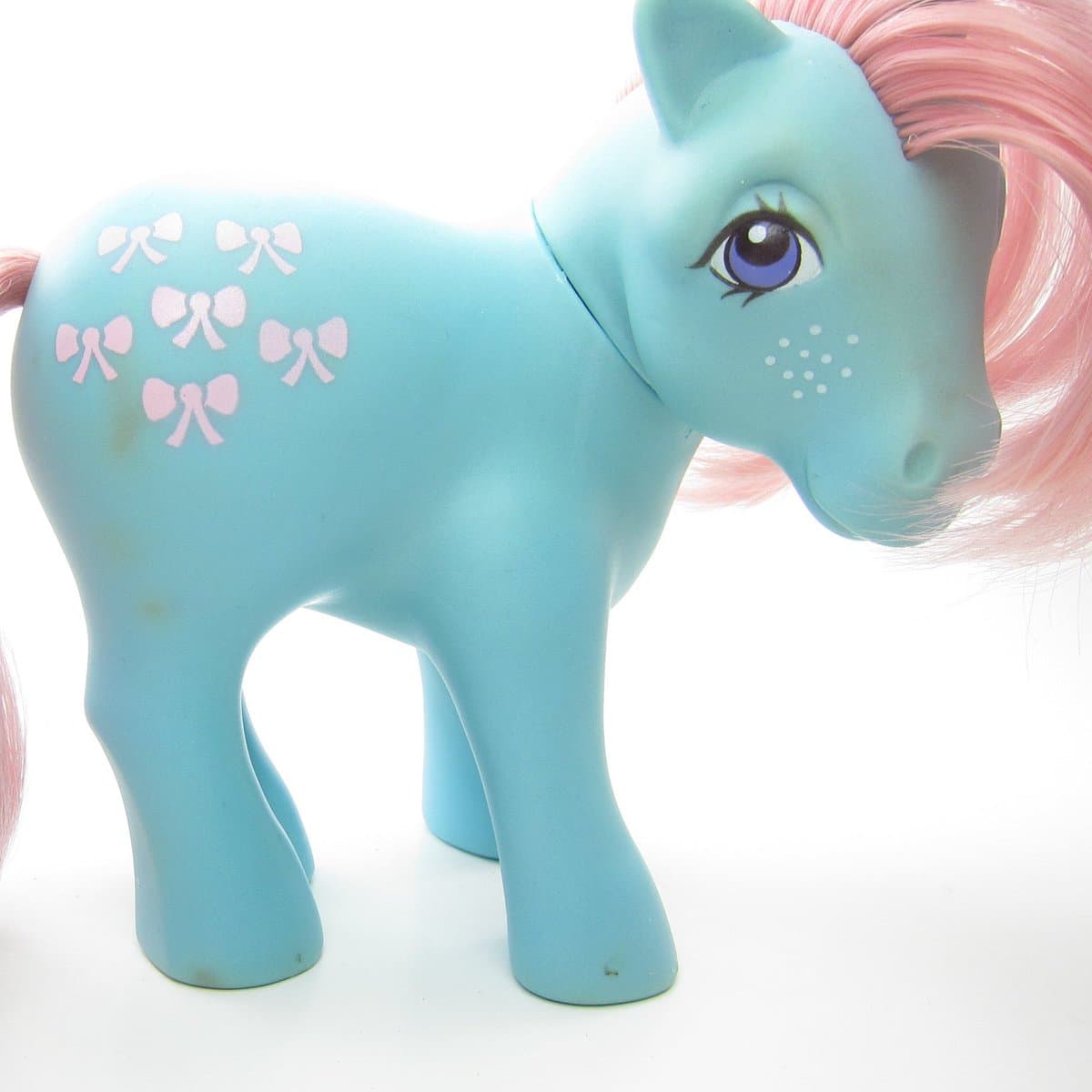 A close-up photograph of a light blue My Little Pony toy with a pink mane and tail, featuring a cutie mark of six pink bows on its flank. The toy is shown standing against a plain white background.