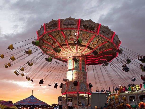A wide shot of a large, illuminated carousel-style amusement park ride with swinging seats against a cloudy sunset sky. The ride features a central column and a large, ornate canopy decorated with red lights, with several empty and occupied seats extending outward on chains. In the background, a blue and orange striped circus tent and other fairground attractions are visible.