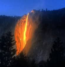 A nighttime photograph of a waterfall illuminated by bright orange light, creating a striking contrast against the dark blue sky and surrounding cliffs. The scene is framed by silhouetted trees in the foreground.