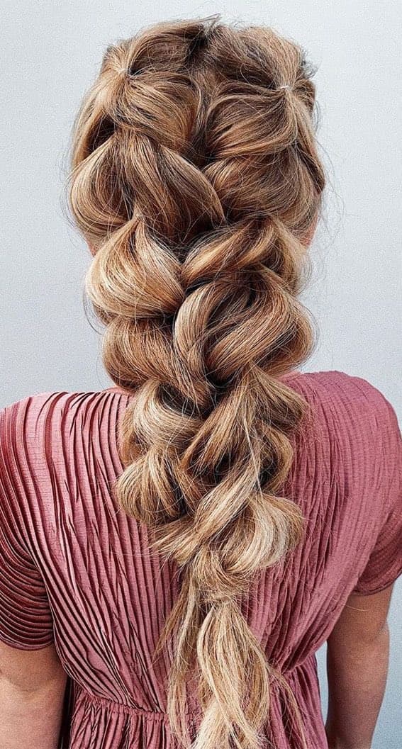 A person is shown from behind, displaying a long, thick, and voluminous blonde braid that falls down their back. They are wearing a short-sleeved, pleated top in a dusty rose color.