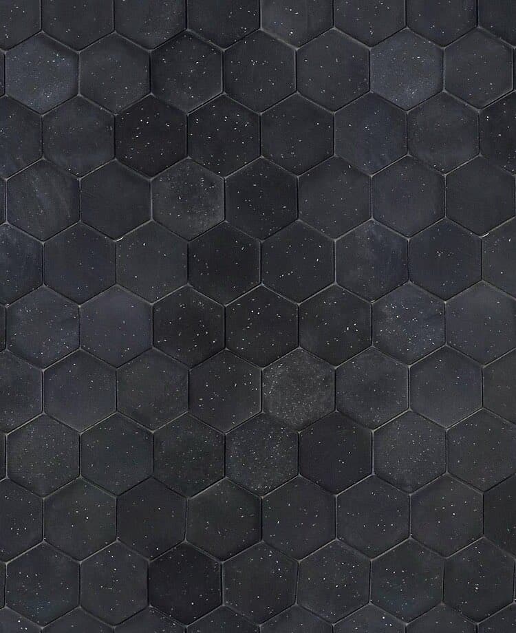 A close-up view of a surface covered in dark, hexagonal tiles arranged in a honeycomb pattern. The tiles have a matte, slightly textured finish with small white speckles scattered across them.