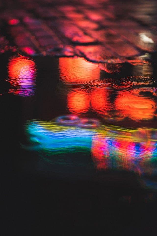 A close-up photograph of colorful neon lights reflecting on a wet, rippling surface, likely a puddle on a street. The image features vibrant streaks of red, orange, blue, and green light distorted by the water's movement against a dark background.