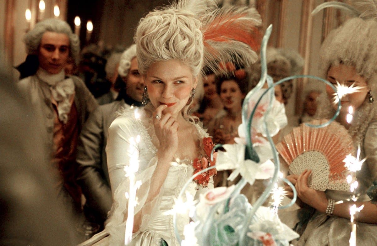 A still from a period film showing a woman in an elaborate 18th-century white gown and powdered wig with feathers, smiling while holding a finger to her lips at a candlelit ball. She is surrounded by other guests in period costumes, including a man in a wig and a woman holding a fan.