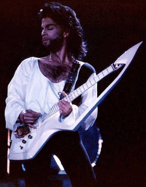 A photograph of a musician with dark hair and a beard, wearing a white shirt, playing a distinctive white V-shaped electric guitar on a stage.
