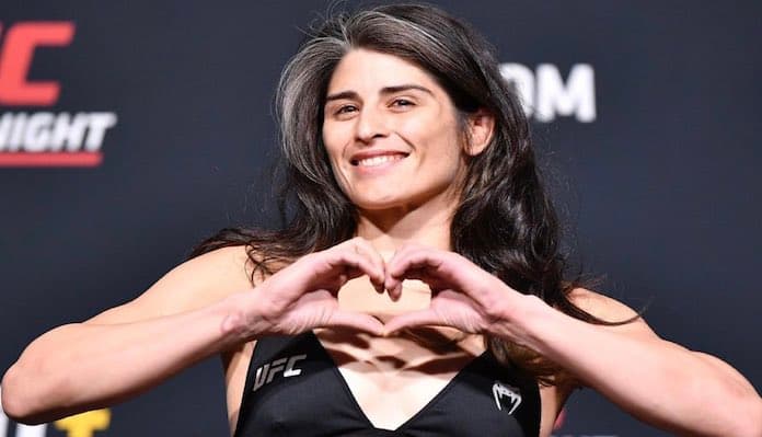 A woman with long dark hair is smiling and making a heart shape with her hands in front of her chest. She is wearing a black top with the UFC logo on it, standing in front of a backdrop featuring UFC branding.