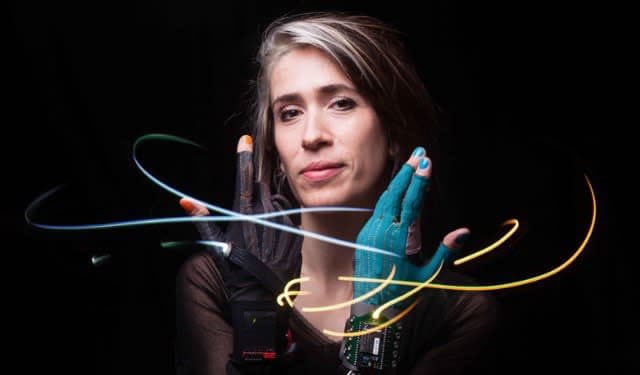 A woman with short, dark hair and a neutral expression is shown against a black background, wearing a dark top and a pair of high-tech, fingerless gloves. The gloves, one black and one teal, are connected to a small electronic device on her chest, and colorful light trails swirl around her hands, suggesting motion or interaction with the technology.
