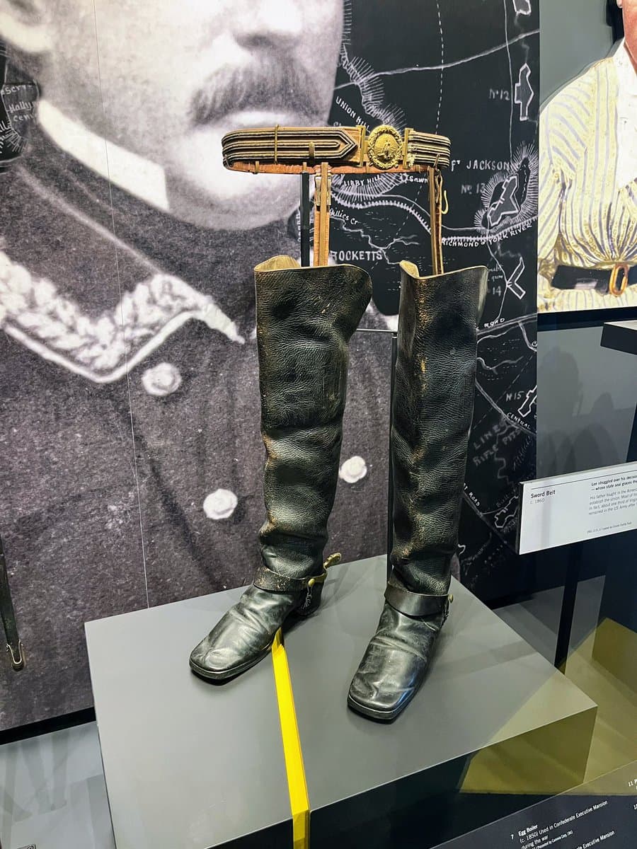 A museum display featuring a pair of tall, worn black leather boots and a brass sword belt mounted on a stand. The background includes a large black-and-white portrait of a bearded man in a military uniform and a historical map.