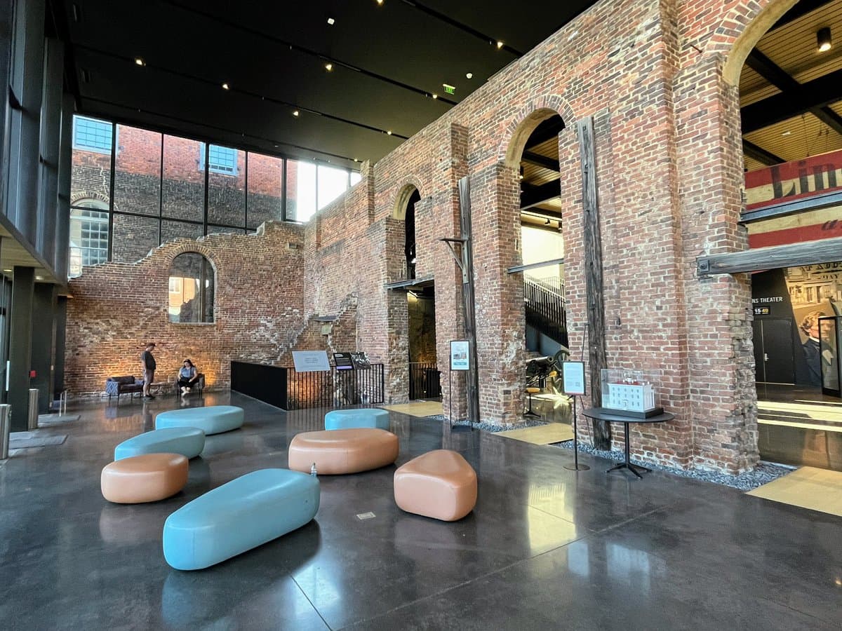 A wide interior shot of a museum space featuring a large, exposed red brick wall with arched openings and wooden support beams. In the foreground, several low, rounded seating cushions in blue and orange are arranged on a polished concrete floor, while a few visitors stand near the back wall.