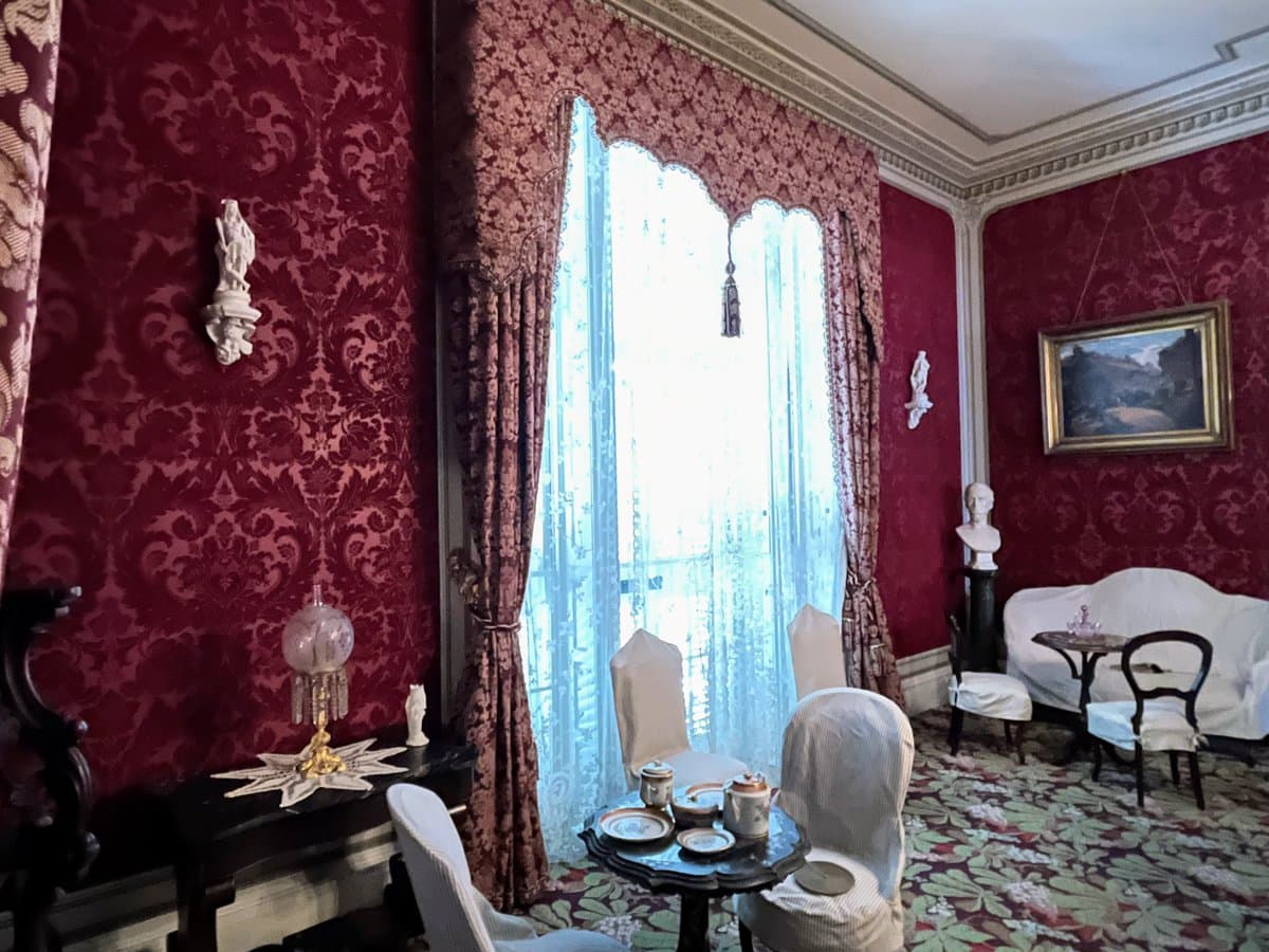 A photograph of an ornate interior room featuring deep red damask wallpaper, white slipcovered furniture, and a large window with patterned drapes. The room is decorated with a white bust on a pedestal, a framed landscape painting, and a small table set with tea service.