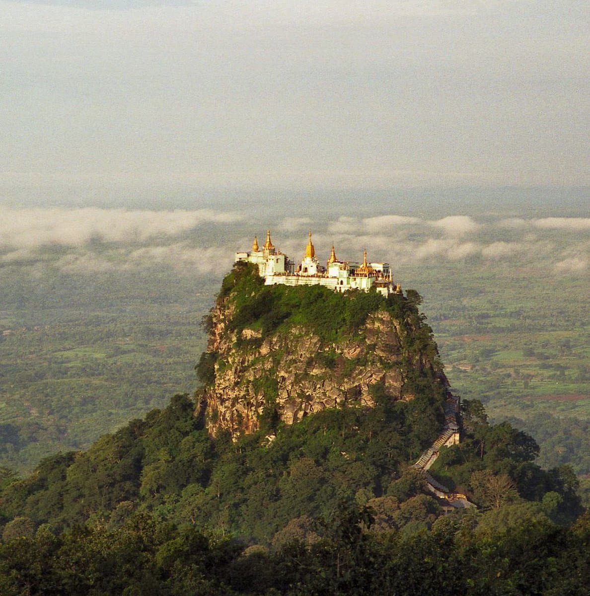 A wide landscape photograph showing a white temple complex with golden spires perched atop a steep, rocky mountain peak. The summit is surrounded by a sea of clouds and a vast, hazy valley below.