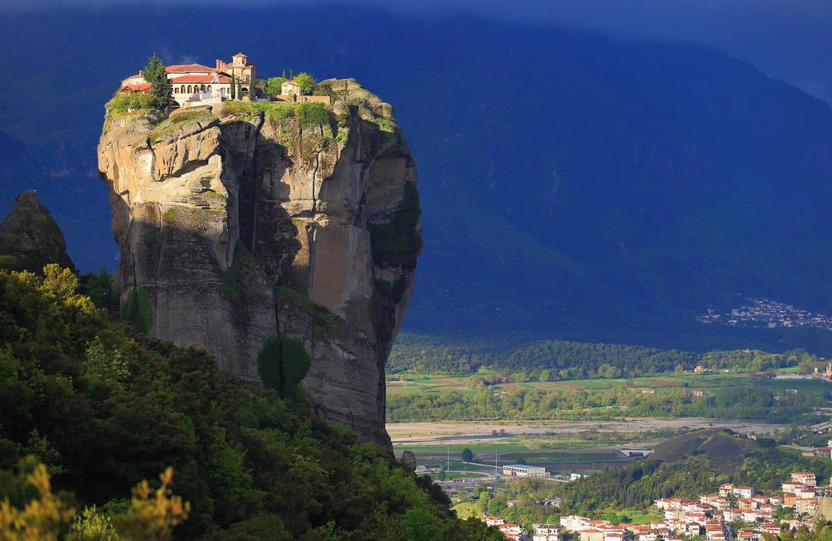 A landscape photograph showing a monastery complex perched atop a massive, towering rock formation, with a green valley and distant mountains in the background.