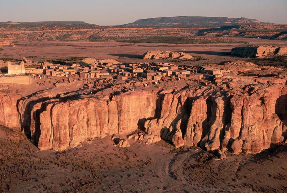 A wide landscape photograph showing a village of flat-roofed, earthen-colored buildings built on the edge of a high, rugged cliff in a desert environment. The foreground features the textured face of the cliff and a dry, rocky valley floor, while the background shows a vast, arid plain stretching toward distant mountains under a clear sky.