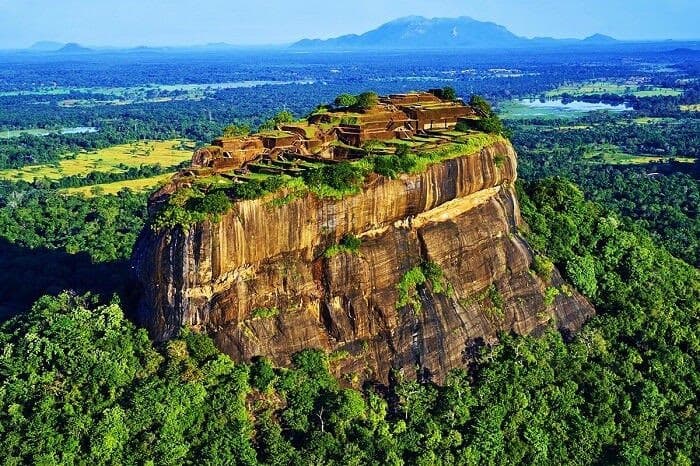 An aerial photograph of a massive, flat-topped rock formation rising above a dense green forest, with ancient brick ruins visible on its summit. In the background, a vast landscape of plains and distant mountains stretches to the horizon under a clear blue sky.