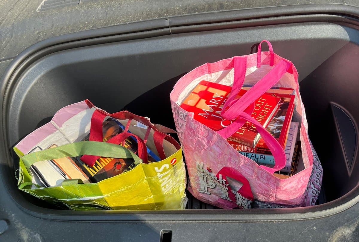 A photograph showing the open trunk of a car filled with two reusable shopping bags overflowing with books. The bags are pink and green, and the books inside include titles such as 'Killing' and 'The Alibi.'