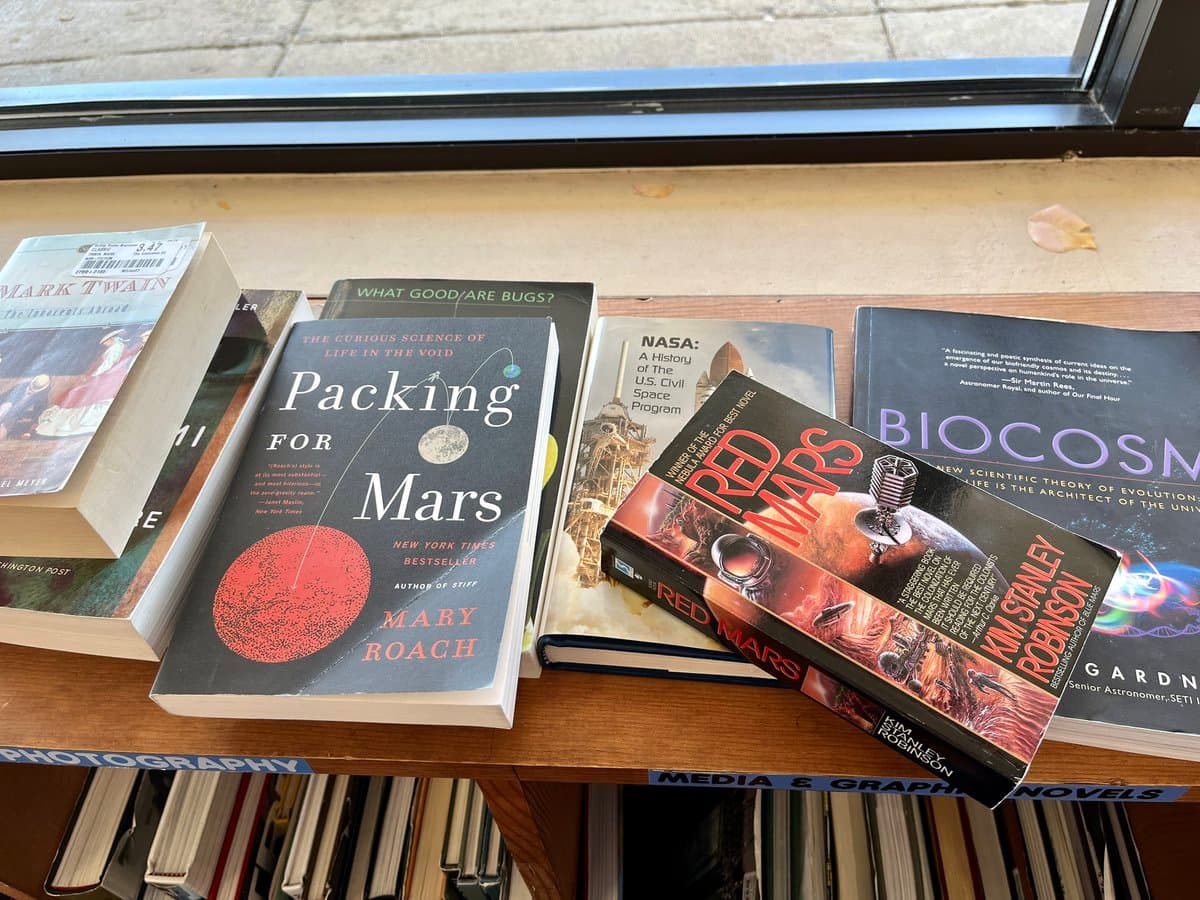 A photograph of several books displayed on a wooden shelf, including titles such as 'Packing for Mars' by Mary Roach, 'Red Mars' by Kim Stanley Robinson, and 'NASA: A History of the U.S. Civil Space Program.'
