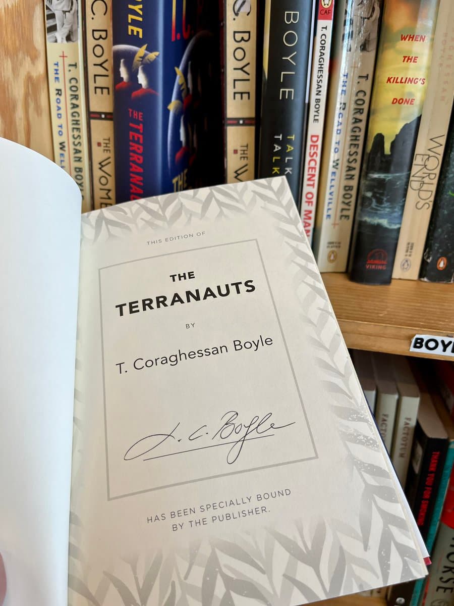 A photograph of an open book displaying the title page for "The Terranauts" by T. Coraghessan Boyle, which features a signature and a note about special binding. The book is held in front of a bookshelf filled with other volumes by the same author.