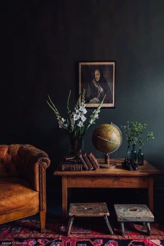 A moody interior photograph featuring a vintage leather armchair, a wooden table with a globe, books, and flowers, and a framed portrait on a dark wall. Two small wooden stools sit on a patterned rug in the foreground. The image has a watermark in the bottom left corner.