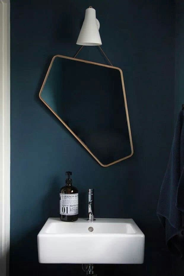 A bathroom interior featuring a dark blue wall, a white wall-mounted sink, and a hexagonal mirror with a wooden frame hanging above it. A bottle of Aesop hand wash sits on the sink next to a chrome faucet.