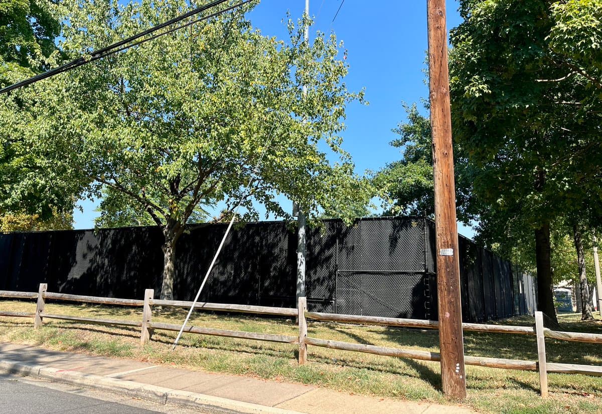 A daytime outdoor photograph showing a black chain-link fence with a privacy screen running along a grassy area behind a wooden split-rail fence. A sidewalk and street are visible in the foreground, with a wooden utility pole and leafy green trees standing against a clear blue sky.