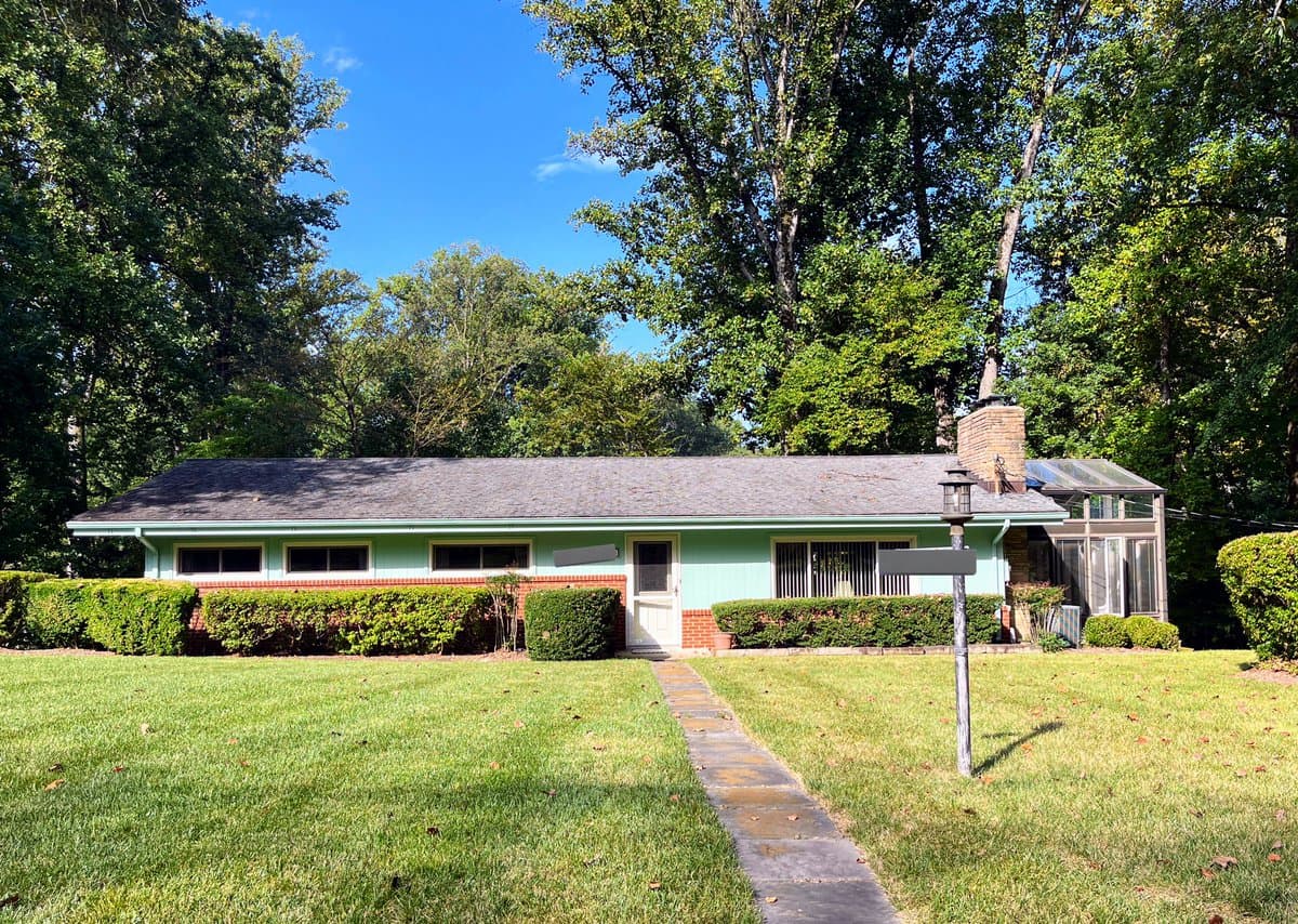 A photograph of a single-story, mint-green house with a brick foundation and a gray shingled roof, set on a grassy lawn with a stone walkway leading to the front door. The house is surrounded by mature trees and hedges, with a screened-in porch visible on the right side.