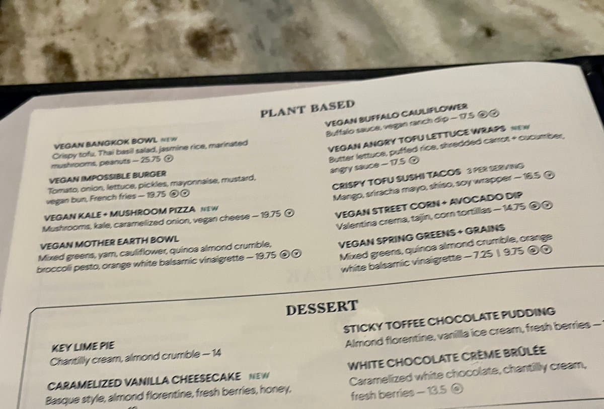 A close-up photograph of a restaurant menu featuring a 'Plant Based' section and a 'Dessert' section. The menu lists various vegan dishes such as the 'Vegan Impossible Burger' and 'Crispy Tofu Sushi Tacos' alongside desserts like 'Key Lime Pie' and 'Sticky Toffee Chocolate Pudding'.