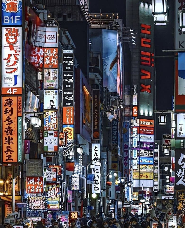 A nighttime street-level photograph of a crowded urban area in Japan, likely Tokyo, featuring a dense array of brightly illuminated vertical neon signs and billboards in Japanese. The scene captures a bustling atmosphere with numerous pedestrians walking below the towering advertisements.